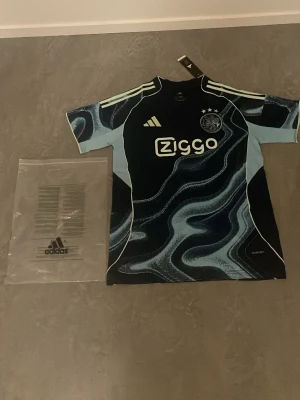 Ajax x Adidas matchtröja blå mönstrad - Säljer en Ajax matchtröja från Adidas i blått med unikt vågigt mönster och ljusblå detaljer. Tröjan har Ziggo-logga på bröstet, klubbmärke och tre stjärnor. Tillverkad i funktionsmaterial, perfekt för fotboll eller träning. Köpte den på Intersport men ville lämna tbx men dem sa att det behövdes kvitto och jag hade tappat bort det och köpte den för 799kr