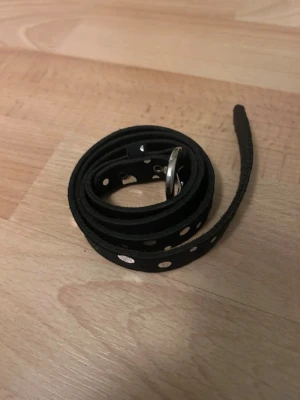 Svart bälte med silverhål - Snyggt smalt svart bälte med silverfärgade hål längs hela bandet och rund metallspänne. Perfekt för att ge en edgy touch till din outfit. Bältet är gjort i skinnimitation och passar till både jeans och kjol.