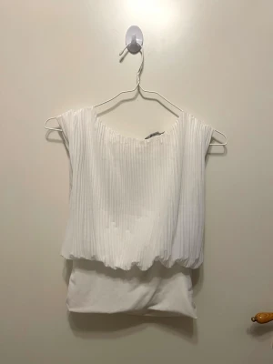 Vit plisserad ärmlös topp - Snygg vit ärmlös topp med plisserad överdel och rund halsringning. Toppen har en knytning i ryggen och ett flowy fall som ger en luftig känsla. Perfekt att styla med jeans eller kjol för en fräsch look.