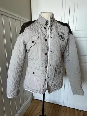 Quiltad beige jacka från Lexington - Quiltad beige jacka från Lexington med mörka axelpartier och tryck på bröstet. Jackan har hög krage, knappar och dragkedja framtill samt flera fickor med lock. Perfekt för dig som gillar klassisk och sportig stil.