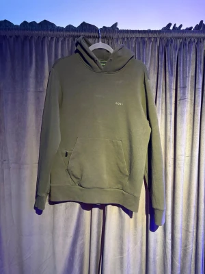 Olivgrön hoodie från BOSS - Säljer en stilren olivgrön hoodie från BOSS med liten logga på bröstet. Hoodien har huva, känguruficka framtill och ribbade muddar vid ärmslut och nederkant. Perfekt för en avslappnad och trendig look.