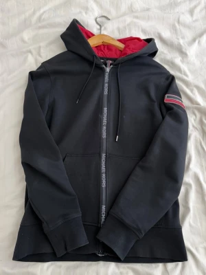 Svart hoodie från Michael Kors - Svart hoodie med dragkedja från Michael Kors. Röd insida på huvan och snygg logga på dragkedjan. Väldigt bra skick och inga fel på den. Köpt från Michael kors butiken. 