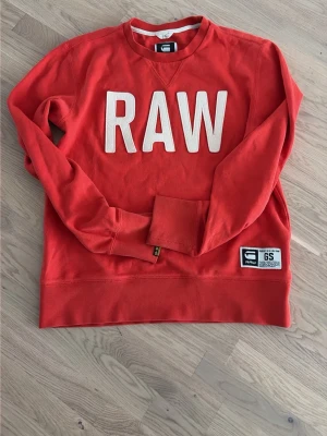 Röd sweatshirt från G-Star RAW - Cool röd sweatshirt från G-Star RAW med stora vita bokstäver 'RAW' på bröstet. Tröjan har rund halsringning, långa ärmar och en patch nertill med logga. Tillverkad i mjukt bomullsmaterial som känns skönt mot huden. Perfekt för en avslappnad streetwear-look.