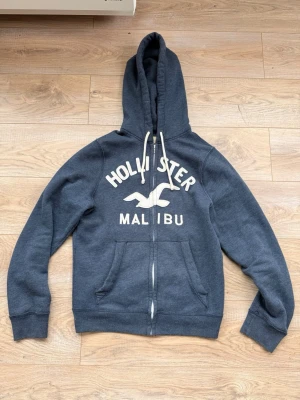 Hollister tröja - Kofta från Hollister med vit broderad logga och texten 'Malibu' på bröstet Storlek M, längd 66-67cm. Tröjan har huva med vita snören, dragkedja framtill och två stora fickor. Mjuk bomullsblandning som är perfekt för en avslappnad stil.