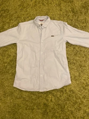 Ljusblå skjorta från Lacoste - Klassisk ljusblå skjorta från Lacoste med button-down krage och vita knappar. Skjortan har lång ärm och den ikoniska krokodilloggan broderad på bröstet. Tillverkad i bomull för en skön och stilren look.