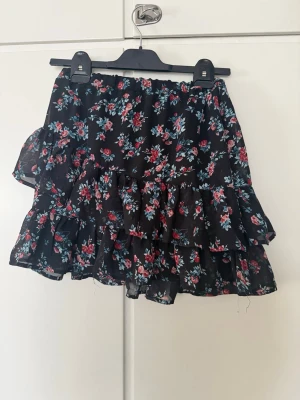 Svart blommig volangkjol - Söt svart kortkjol med blommigt mönster i rött, blått och vitt. Kjolen har flera volanger som ger en flowy känsla och elastisk midja för skön passform. Perfekt för dig som gillar romantisk och trendig stil.
