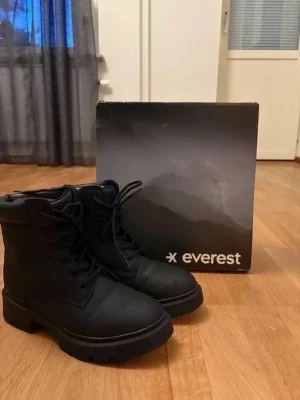 Svarta kängor från Everest - Stilrena svarta kängor från Everest med grov sula och snörning framtill. Kängorna är tillverkade i ett tåligt material och har en klassisk, robust design som passar perfekt till höst och vinter. Vattentäta och med bekväm vadderad kant upptill.