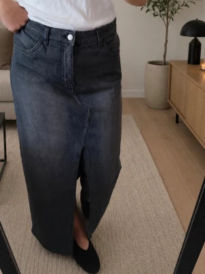 Grå  lång jeanskjol från Kappahl - Säljer en grå lång jeanskjol från Kappahl med snygg slits framtill. Kjolen har klassiska jeansdetaljer som fickor och knappstängning. Perfekt för dig som gillar en stilren och trendig look.