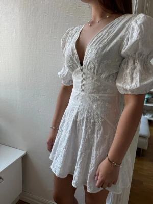 Adoore Vit  - Kort vit klänning med puffärmar, djup v-ringning och knappar framtill. Perfekt inför sommaren eller exam! Endast använd en gång på skolavslutning💗💗