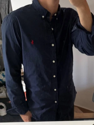 Mörkblå skjorta från Ralph Lauren - Klassisk mörkblå skjorta från Ralph Lauren med knappslå och vit knappdetalj. Skjortan har lång ärm, button-down krage och den ikoniska röda logotypen broderad på bröstet. Tillverkad i mjuk bomull för en bekväm känsla. Lite missfärgad annars bra skick 