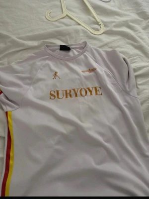 Vit Suryoye sporttröja - Säljer en vit sporttröja med texten SURYOYE i guld på bröstet. Tröjan har röda och gula ränder längs sidorna och detaljer i guld, inklusive en liten figur och en symbol på bröstet. Perfekt för träning eller match.