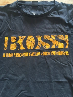 Svart BOSS t-shirt med gul logga - Snygg svart t-shirt från BOSS med gul logga och mönstrad text framtill. Klassisk rund halsringning och korta ärmar. Perfekt för dig som vill ha en statement-tee med coolt tryck.
