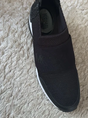 Svarta slip-on sneakers Michael Kors - Michael Kors sneakers – storlek 38  Snygga och bekväma svarta slip-on sneakers från Michael Kors med vita sulor och fina glittriga detaljer baktill. Stilren design som passar till både vardag och finare tillfällen. Säljs eftersom de inte kommer till användning. Hör gärna av dig vid intresse eller frågor! 😊