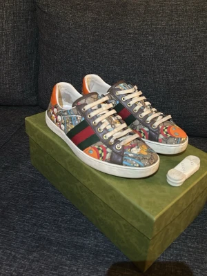 Gucci sneakers med färgglatt mönster - (ÄKTA)Kan skicka fler bilder privat. Pris kan diskuteras. Unika Gucci sneakers med färgglatt grafiskt mönster, klassiska gröna och röda sidoremsor samt orange detaljer vid hälen. Skorna har vita snören och sula, och är tillverkade i läder och textil. Perfekta för dig som vill sticka ut med din stil.