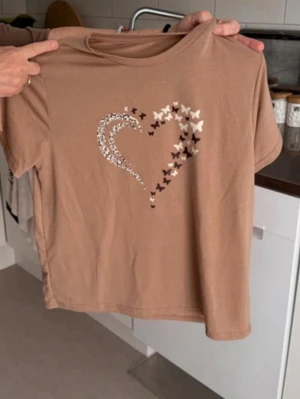 Beige t-shirt med hjärtdetalj - Säljer en beige t-shirt med korta ärmar och ett snyggt hjärtmotiv framtill, dekorerat med små fjärilar i olika färger. Perfekt för dig som gillar unika detaljer och en avslappnad stil. Rund halsringning och mjukt material.