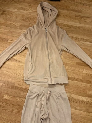 Beige mjukisdress  - Säljer en beige mjukisdress i velour med dragkedja framtill och huva. Setet består av en hoodie med fickor och matchande byxor med dragsko i midjan. Perfekt för chill dagar hemma eller när du vill vara extra bekväm.