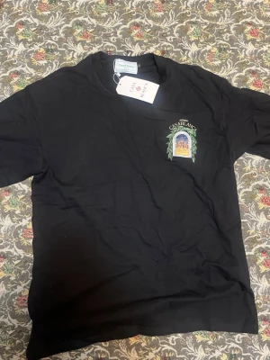 Svart t-shirt från Casablanca - Snygg svart t-shirt från Casablanca med tryck på bröstet som föreställer en byggnad inramad av gröna blad och texten 'Avenida Casablanca'. Klassisk passform och rund hals. Perfekt för dig som vill ha en stilren men unik look.