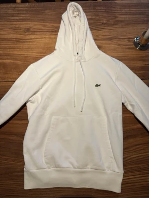Vit hoodie från Lacoste med logga - Säljer en stilren vit hoodie från Lacoste med klassisk krokodillogga på bröstet. Hoodien har huva med dragsko och en stor magficka framtill. Tillverkad i mjukt bomullsmaterial som känns skönt mot huden. Perfekt för en avslappnad och trendig look.