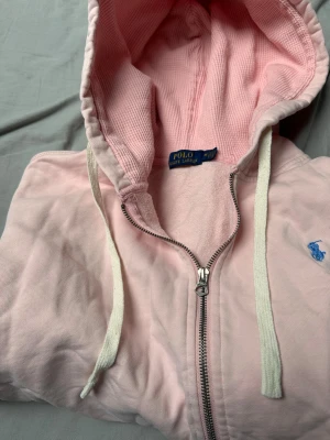 Ljusrosa hoodie från Polo Ralph Lauren - Snygg ljusrosa hoodie från Polo Ralph Lauren med dragkedja framtill och vit dragsko i huvan. Tillverkad i mjuk bomull med blå broderad logga på bröstet. Perfekt för en chill och avslappnad stil.