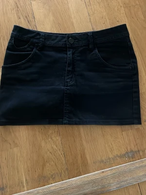 Svart jeanskjol - Snygg svart kortkjol i denim-look med fram- och bakfickor samt hällor för bälte. Kjolen har en klassisk knappstängning framtill och raka linjer för en clean stil. Perfekt att matcha med en crop top eller hoodie. Midjemått är 38cm rakt över!