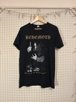Svart Behemoth band t-shirt - Säljer en ovanlig svart t-shirt med Behemoth-tryck fram och texten 'The Ceremony of Wolves Pagan Black Metal' på ryggen. T-shirten har korta ärmar, rund hals och är gjord i mjuk bomull. Perfekt för dig som gillar metal och vill ha en unik bandtröja.