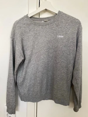 Grå sweatshirt med Love-tryck - En basic grå sweatshirt med rund halsringning och diskret vitt 'Love'-tryck på bröstet. Tröjan har långa ärmar, ribbade muddar och är gjord i mjukt sweatshirtmaterial. Perfekt för en avslappnad och stilren look.