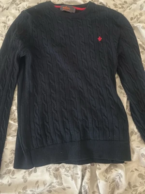 Mörkblå kabelstickad tröja Morris -  mörkblå kabelstickad tröja från Morris med rund halsringning och röd broderad logga på bröstet. Tillverkad i 100% lammull för extra mjuk och varm känsla. Lite liten i storlek skulle jag säga men storlek är s/xs! ☺️