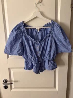 Blåvit randig blus från H&M - Säljer en blå blus med vita ränder från H&M. Blusen har korta ärmar, knappar framtill och en rynkad, elastisk midja som ger en snygg passform. Materialet är lätt och luftig bomull, perfekt för varma dagar. Rundad halsringning med volangdetaljer.