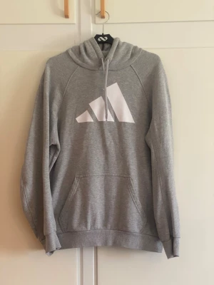 Grå hoodie från Adidas med logga - Snygg grå hoodie från Adidas med stor vit logga på bröstet. Klassisk känguruficka framtill, ribbade muddar och justerbar huva med dragsko. Tillverkad i mjukt sweatshirtmaterial som är perfekt för chill eller träning.