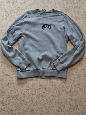 Ljusblå sweatshirt från EA7 - Ljusblå sweatshirt från EA7 Emporio Armani med rund halsringning och ribbade muddar vid ärmslut och nederkant. Tröjan har en broderad EA7-logga på bröstet och är tillverkad i mjuk bomull. Perfekt för en avslappnad och stilren look.