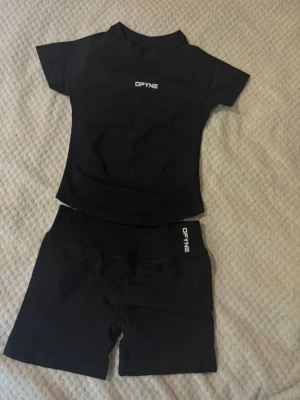 Svart träningsset från DFYNE - Säljer ett svart träningsset från DFYNE med en tight t-shirt och matchande shorts. Båda plaggen har vit DFYNE-logga framtill och baktill. Setet är gjort i ett stretchigt och mjukt material som passar perfekt för gym eller löpning. Storlek S