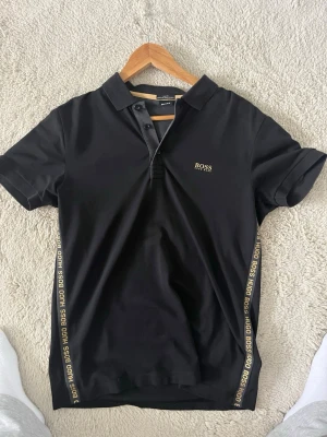 Svart pikétröja från Hugo Boss - Snygg svart pikétröja från Hugo Boss med guldfärgade logotexter längs sidorna och liten logga på bröstet. Klassisk krage och korta ärmar. Perfekt för en clean och stilren look.