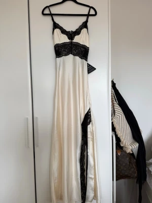 Champagnefärgad långklänning med spets - Elegant långklänning i champagnefärgad satin med svarta spetsdetaljer. Klänningen har tunna axelband, öppen rygg och vackra spetsinslag både fram och bak. Materialet ger en lyxig känsla och den figurnära passformen framhäver siluetten.