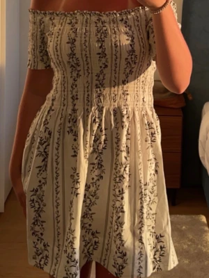 Offshoulder blommig kortklänning - Söt offshoulder kortklänning i vitt med svart blommigt mönster. Klänningen har smockad överdel och ärmar som sitter nedanför axlarna. Jättefin på sommaren💕