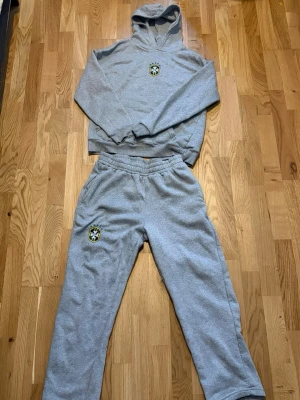 Grå hoodie med Brasilien-märke - Mysigt grått loungewear-set med hoodie och matchande byxor. Hoodien har känguruficka, huva och ett Brasilien-inspirerat märke på bröstet. Byxorna har resår i midjan och samma märke på benet. Perfekt för chill dagar hemma eller när du vill vara bekväm.