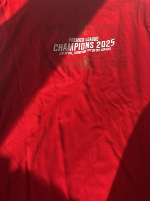 Liverpool FC Champions 2025 T-shirt - Röd t-shirt från Liverpool FC med trycket 'Premier League Champions 2025' och klubbens logga på bröstet. Klassisk rund hals och korta ärmar. Tillverkad i mjuk bomull, perfekt för alla fans som vill visa sitt stöd för laget.