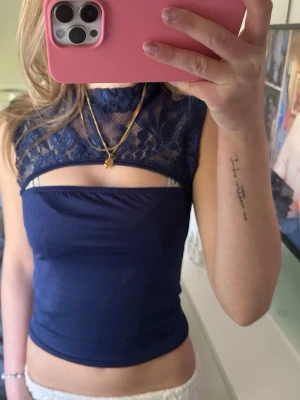 Marinblå spets topp med cut out - Trendig marinblå topp med spetsdetaljer upptill och en snygg cut out över bröstet. Hög hals och ärmlös design ger en modern look. 
