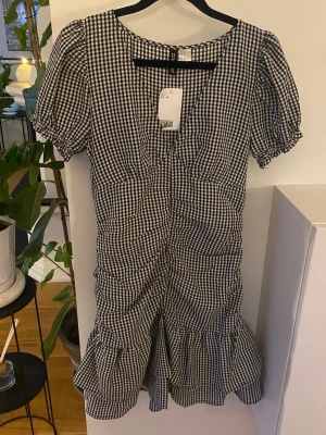 Kort svart/vit rutig klänning M från H&M Divided Slimfit - En kort klänning i vävd kvalitet. Klänningen har v-ringning och avskärning under bysten samt kort puffärm med klädd resår och volangkant vid ärmslut. Rynkat, figurnära liv med draperad effekt samt kjol med avskärning och dubbla volanger nedtill. Dold dragkedja i ena sidan. Fodrad.  Ny med prislapp och slutsåld på H&M  Axel: Bredd: 33.7 cm (storlek 38) Baksida: Längd: 84.5 cm (storlek 38)  Slimfit  