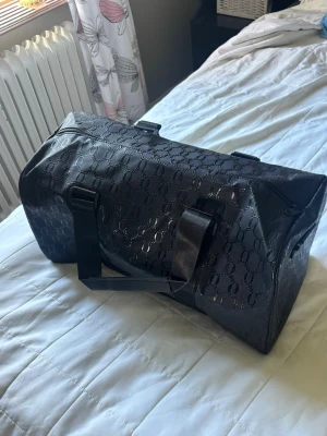 Svart weekendbag med mönster - Snygg svart weekendbag med diskret mönster i material som liknar konstläder. Väskan har två robusta handtag och dragkedja upptill. Perfekt storlek för resor eller gymmet och ger en stilren look.