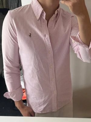 Rosa och vit randig skjorta Polo Ralph Lauren - Klassisk rosa och vit randig skjorta från Polo Ralph Lauren med button-down krage och broderad logga på bröstet. Skjortan har lång ärm, normal passform och knäppning framtill. Perfekt till jeans eller chinos för en clean och stilren look.