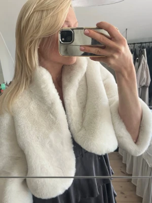 Vit  fuskpälsbolero - Supersnygg vit fuskpälsväst med bred krage och kortare modell. Västen har en mjuk och lyxig känsla, perfekt att slänga över en klänning för extra stilpoäng. Ingen synlig knäppning framtill och rundad nederkant. Helt ny!