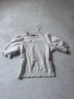 Vit stickad tröja med puffärm - Snygg vit stickad tröja med puffärmar och rund hals helt ok nopprig men kan lösas lätt med ex rakhyvel. Tröjan har ribbade muddar vid ärmslut och nederkant. Materialet är mjukt och ser ut att vara en ull- eller ullmix. Perfekt för dig som gillar trendiga detaljer och en clean look.