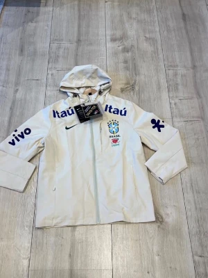 Vit Brasil Nike vindjacka med huva - Vit vindjacka från Nike med Brasil-logga och flera sponsortryck i blått. Jackan har huva med dragsko, hel dragkedja och är tillverkad i GORE-TEX-material. Perfekt för dig som gillar sportiga och funktionella plagg.