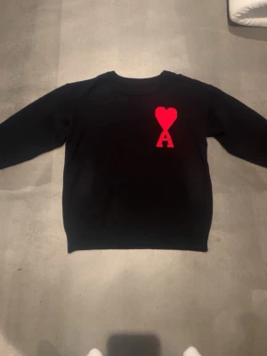 Svart stickad tröja Ami Paris - Snygg svart stickad tröja från Ami Paris med deras ikoniska röda hjärta och bokstaven A broderat på bröstet. Tröjan har rund halsringning och långa ärmar. Perfekt för dig som gillar stilrena plagg med en twist.