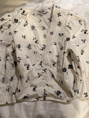 Vit blommig blus med puffärm - Supersöt vit blus från Vintage Stories med svarta blommönster, puffiga långärmar och knappar framtill. V-ringad och figursydd modell. Perfekt till jeans eller kjol. Aldrig använd. Tveka inte att höra av er💝😽