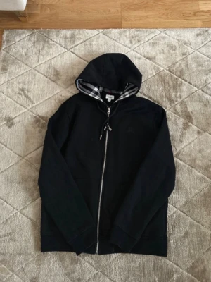 Svart hoodie  - Säljer en svart hoodie med dragkedja och klassiskt rutigt foder i huvan. Hoodien har dragsko i huvan och ribbade muddar vid ärmslut och nederkant. Perfekt för dig som gillar stilrena plagg med en twist. Den är Xl men passar som L-M