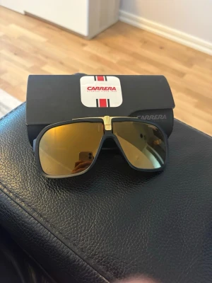 Carrera solglasögon - Snygga svarta Carrera solglasögon med spegelglas i guld och cool detalj i guld på bryggan. Klassisk pilotformad modell som ger en sportig vibe. Kommer med originalfodral från Carrera.