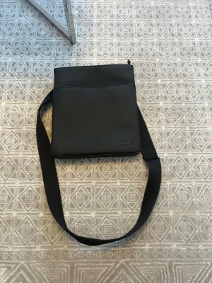 Lacoste väska - Snygg svart axelväska från Lacoste med minimalistisk design och diskret krokodillogga i hörnet. Väskan har justerbar axelrem i tyg och är tillverkad i ett slätt material som ger en clean look. Perfekt för dig som vill ha en stilren och praktisk väska.