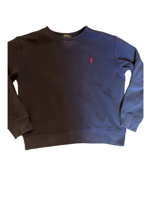 Mörkblå sweatshirt från Polo Ralph Lauren - Klassisk mörkblå sweatshirt från Polo Ralph Lauren med rund halsringning och ribbade muddar. Tröjan har den ikoniska röda logotypen broderad på bröstet. Perfekt för en avslappnad och stilren look. Storlek 14-16 år 
