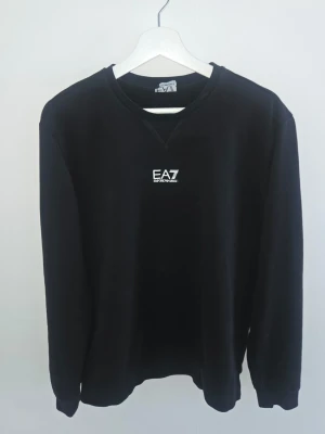 Svart sweatshirt från EA7 Emporio Armani - Stilren svart sweatshirt från EA7 Emporio Armani med vitt logotryck på bröstet. Klassisk rund halsringning och ribbade muddar vid ärmslut och nederkant. Perfekt till ett avslappnat streetwear-look.gott skick se bilder, Storlek L passar M/S bättre
